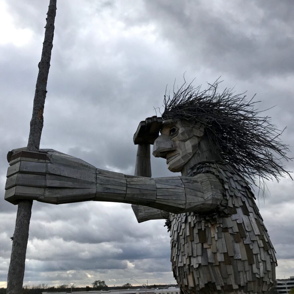 A wooden sculpture from the Morton Aboretum of a person holding a staff and their other hand held flat above their eyes as if they are looking off into the distance