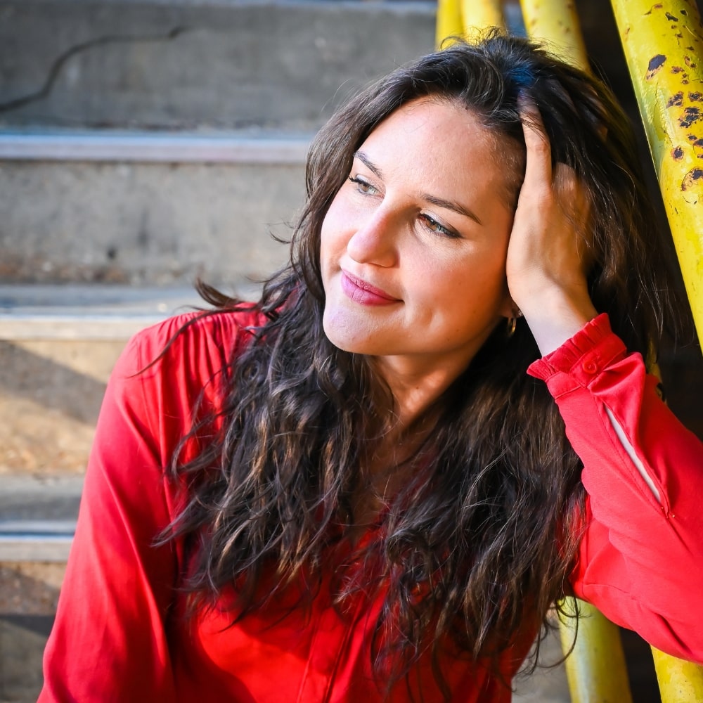 Ashlee sitting on concrete stairs leaning against the yellow railing with her arm and that hand holding head as she looks off the the left of photo. She's smiling and wearing a red long sleeve shirt with her long brown hair covering her shoudlers.