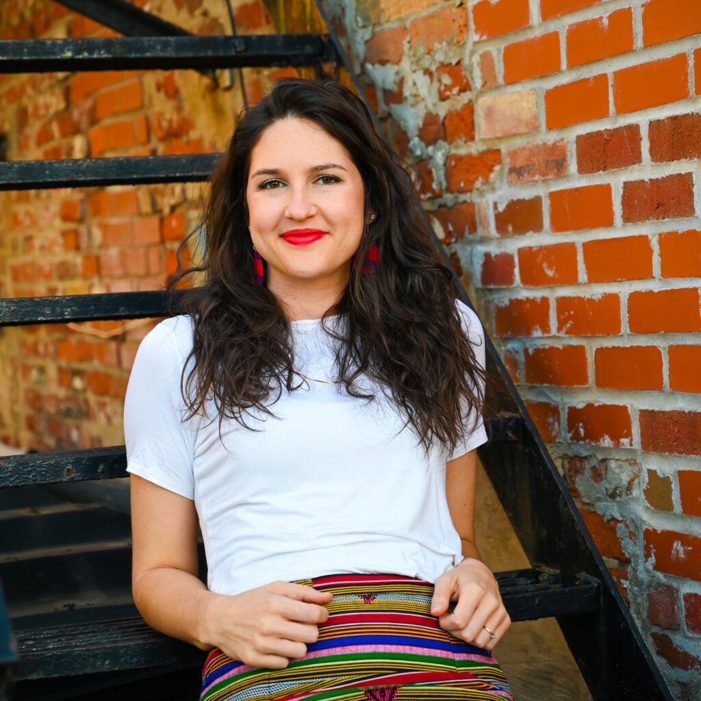 Ashlee Sang Brand Consultant is sitting on a black staircase leaning back with her elbows leaning on the step behind her. The stairs go up next to a brick wall. She is wearing a white t-shirt and colorful striped bottoms with a bright red shade of lipstick, smiling at the camera. The image shape is that of a rounded X.