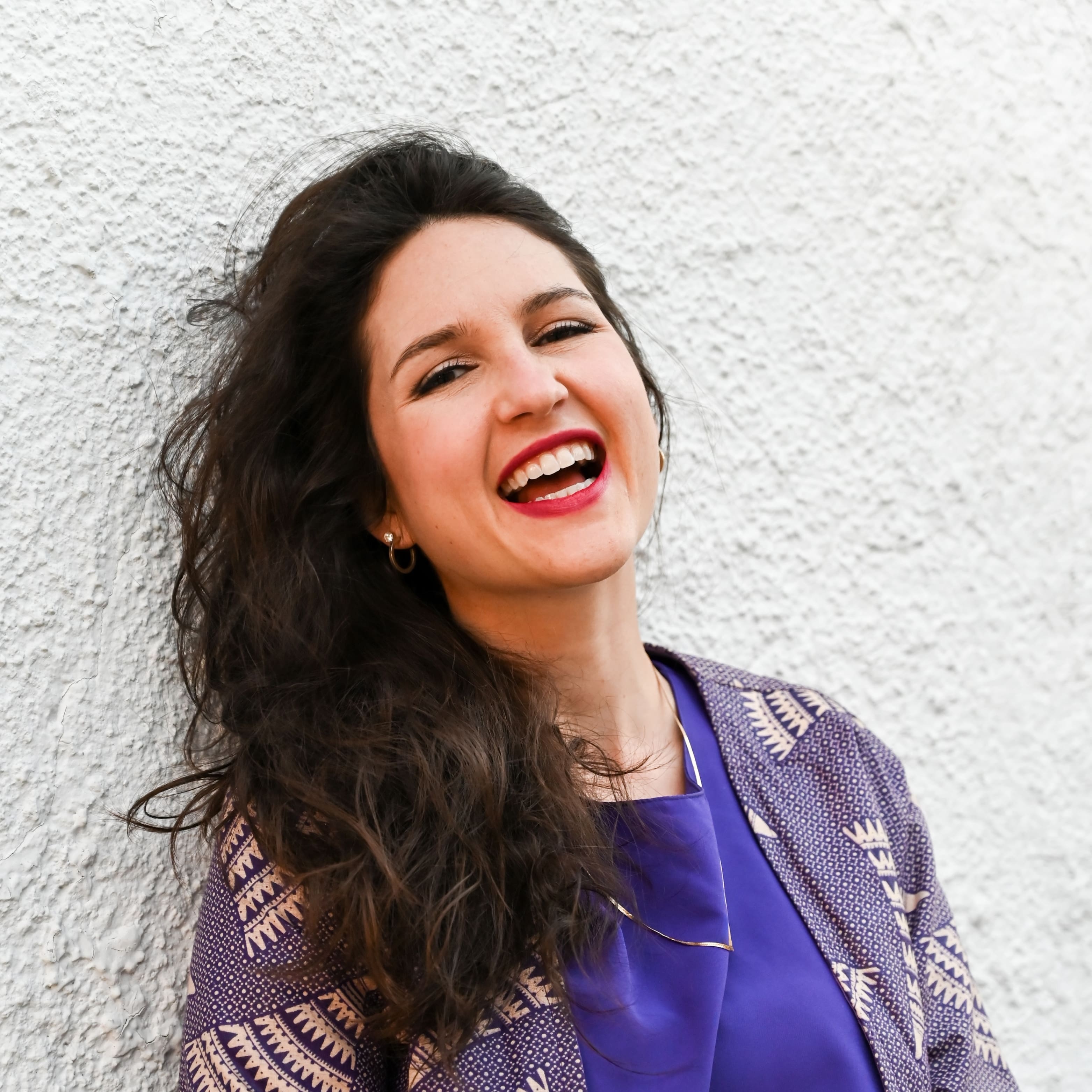 Ashlee looks so happy as she appears to be laughing while leaning against a while wall in a light purple jacket and purple top. The shape of the image is a rounded X.