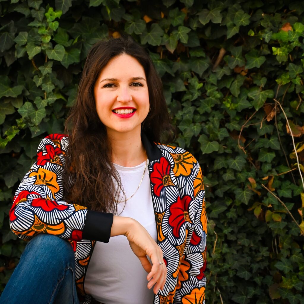 Ashlee Sang Brand Consultant sitting in front of any ivy covered wall. She has one leg bent up with her arm resting on it, smiling at the camera in a colorful jacket.