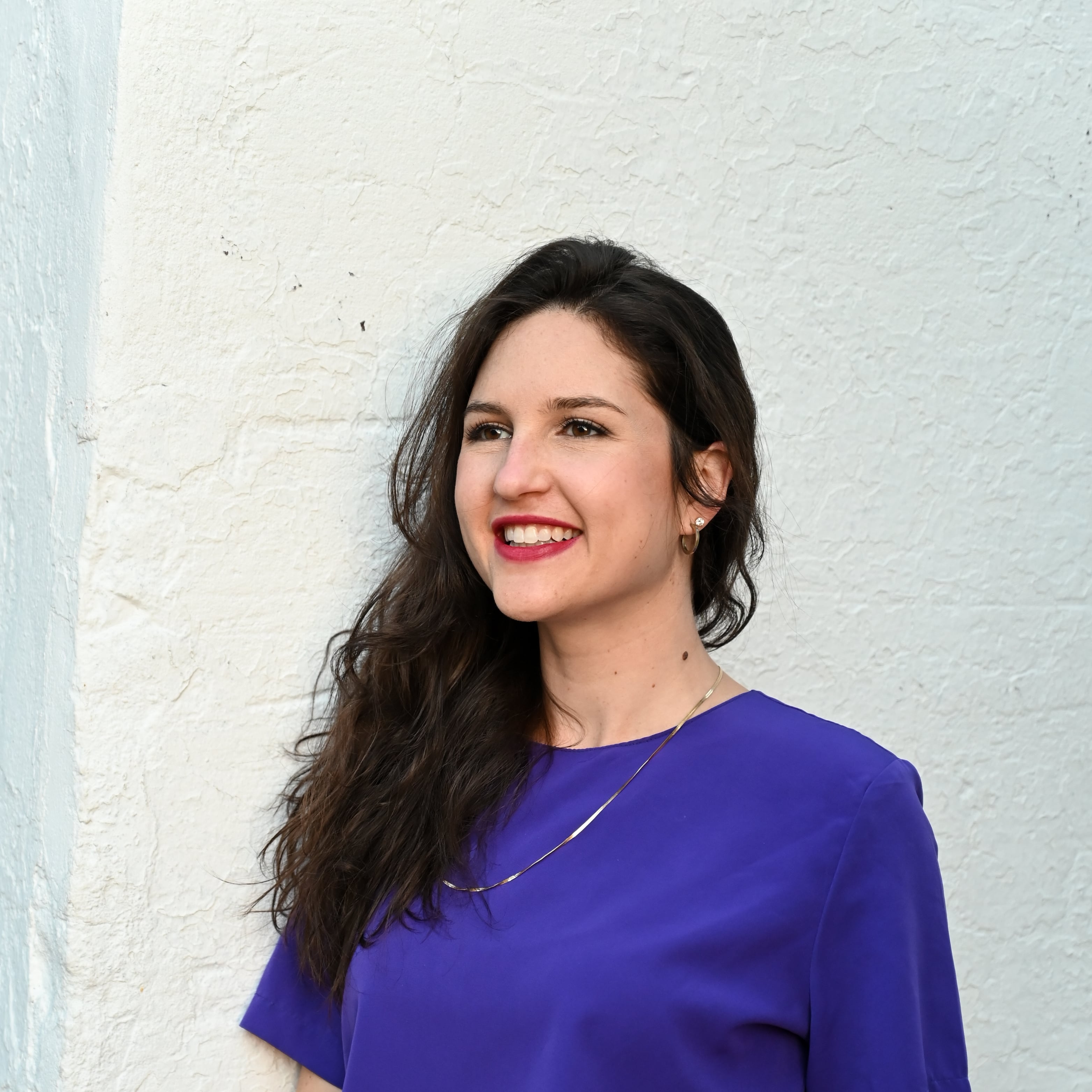 Ashlee Sang Brand Expert is standing in front of a white wall wearing a purple t-shirt. Her long brown hair is down over one shoulder as she smiles in red lipstick looking off to the left. The shape of the image is a rounded X