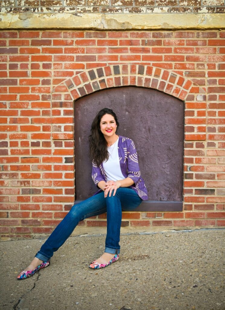 Ashlee sitting on the window sill of a nicely boarded up arched window opening in a brick building.