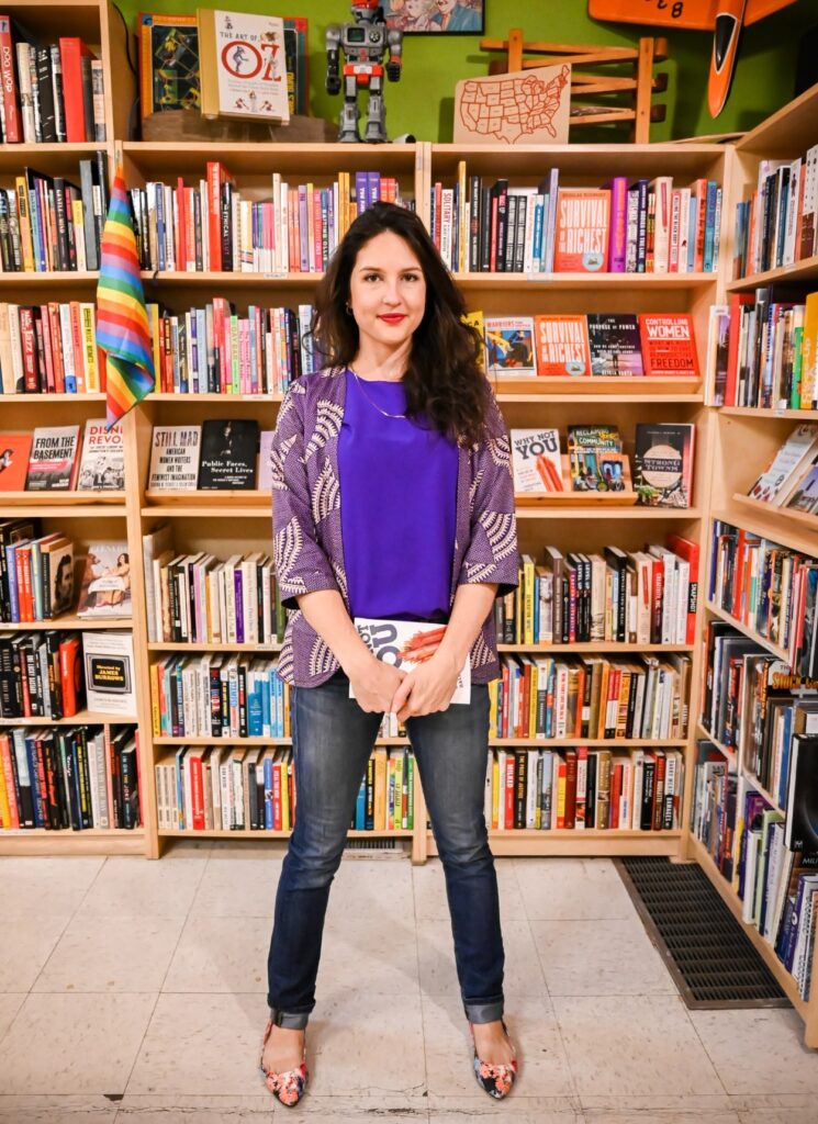 Podcast host Ashlee Sang is in front a book shelf full of books in a book store. She is holding a book with both hands down in front of her and her feet are spread shoulder width apart in a confident stance.