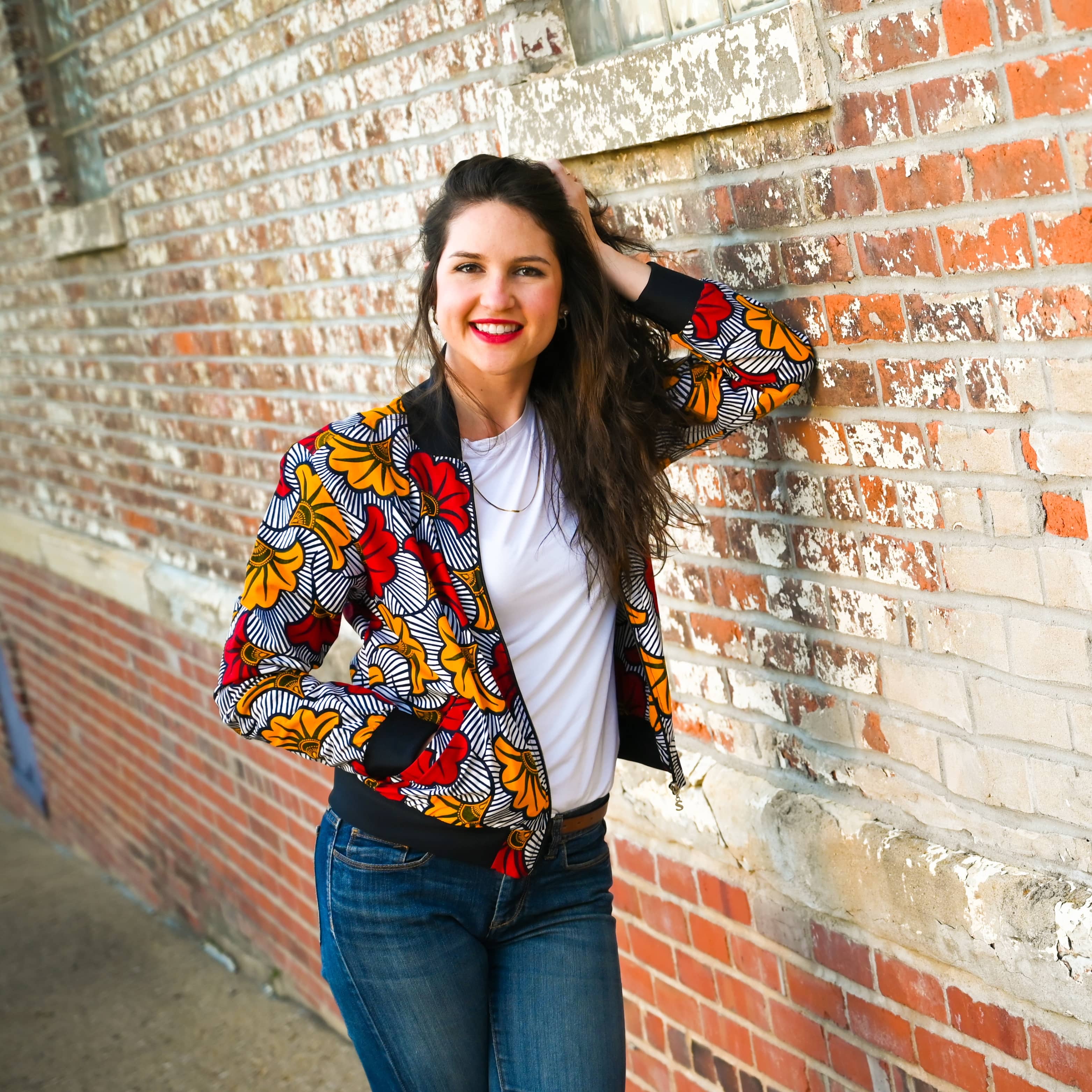 Ashlee looking cool leaning on a brick wall with her elbow as her arm is bent up with her hand in her hair, the other in her colorful jacket pocket. She smiles at the camera.