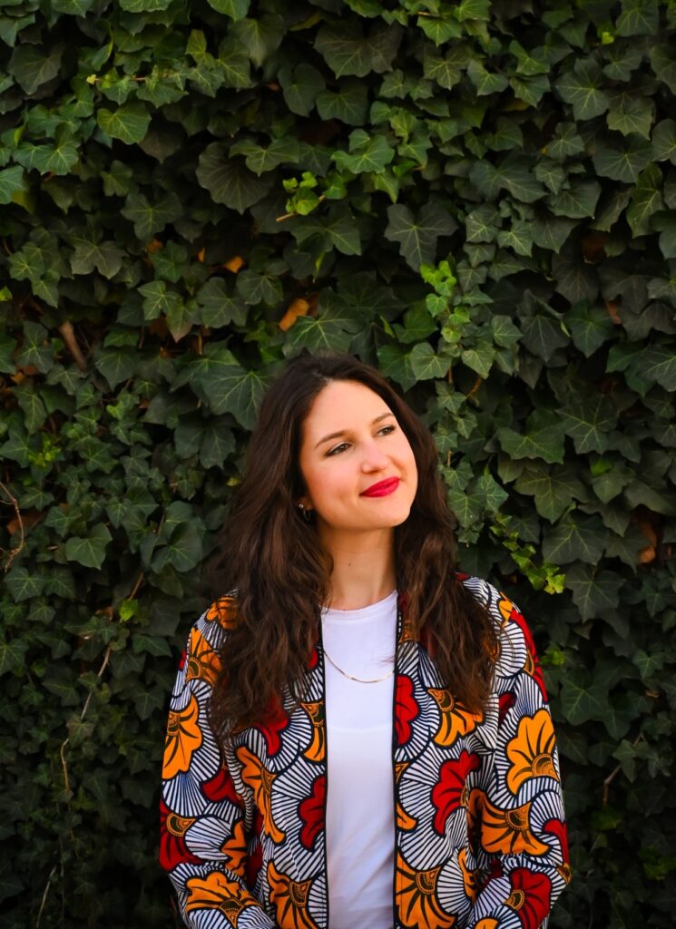 Ashlee Sang standing in front of an ivy-covered wall in a colorful jacket looking off to the side with a smile.