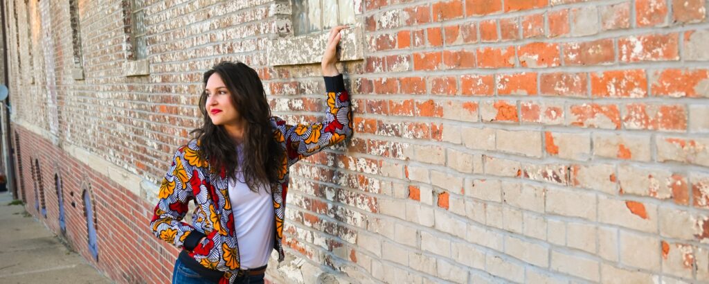Ashlee Sang Brand Consultant poses next to a brick wall, leaning with one arm on the wall and the other hand in her colorful jacket's pocket. She's looking away from the wall to left in the photo. The shape of the image is an upside down half circle.
