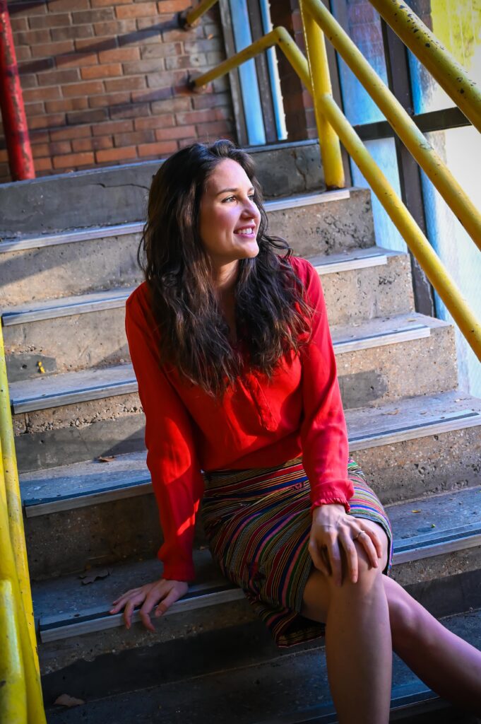 Ashlee Sang Brand expert is sitting on concrete stairs with yellow metal rails. She's wearing a long sleeve red top and colorful striped skirt. She's sitting with her legs crossed, one hand on her knee and the other on the stair, looking off to the right and smiling.